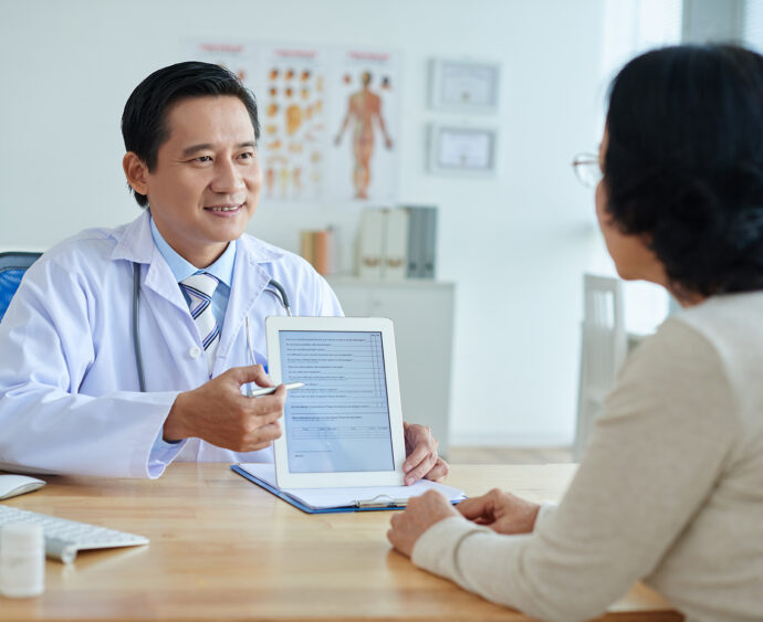 Middle-aged Asian physician looking at female patient with smile while pointing at screen of digital tablet, waist-up portrait